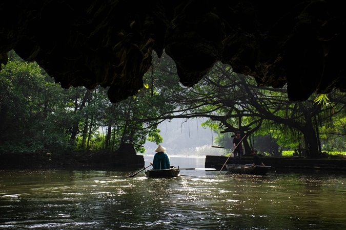 Un vietnamita fotografato di spalle mentre pagaia sulla sua piroga in legno tradizionale immerso nella penombra di un paesaggio fluviale.