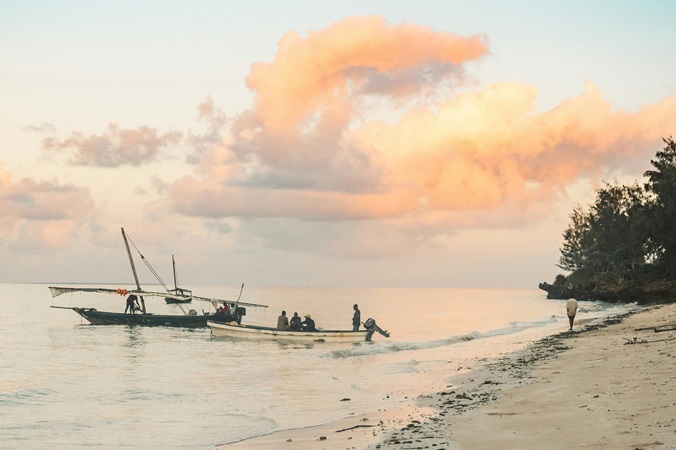 Scorcio di una spiaggia di Zanzibar, al tramonto, con una barca tradizionale e una barca a motore accostate e un uomo che cammina sul bagnasciuga.