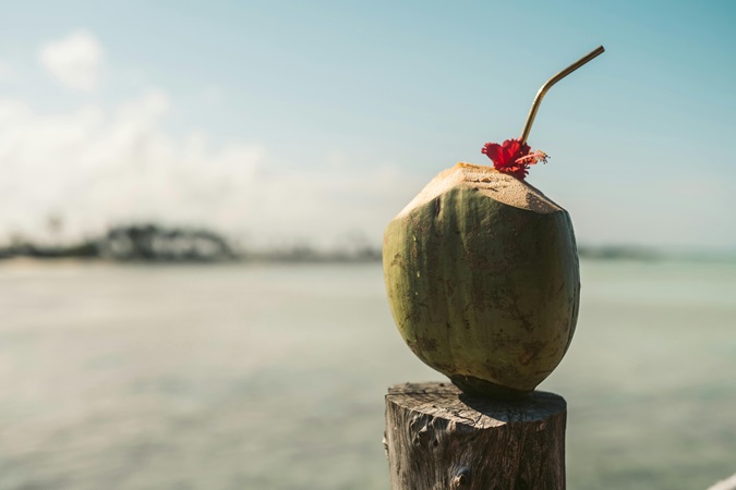 Un cocktail offerto in una noce di cocco, poggiata sul palo di legno di un molo. Sullo sfondo il paesaggio marino di Zanzibar: una tranquillità che non si vuole rovinare con problemi di documenti, per questo proponiamo una guida completa per la richiesta del visto e l'assicurazione per Zanzibar.